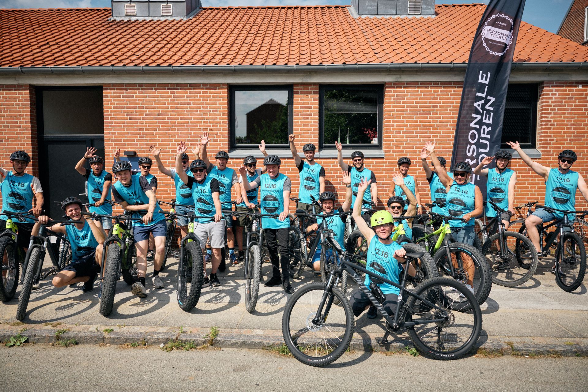 En gruppe mennesker poserer til et billede med deres cykler foran en murstensbygning.