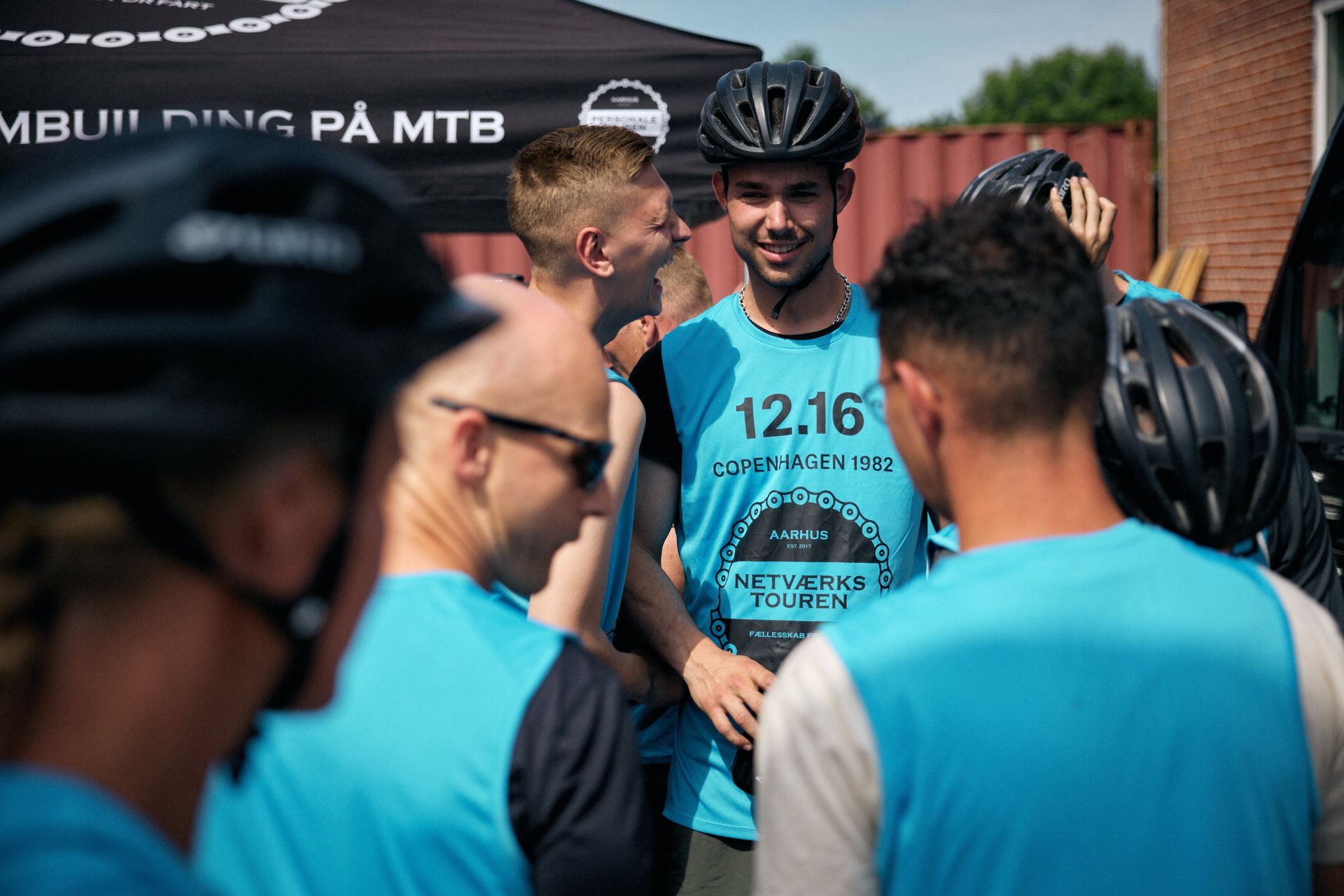 Cyklister i blå veste og hjelme, smilende og taler udendørs.