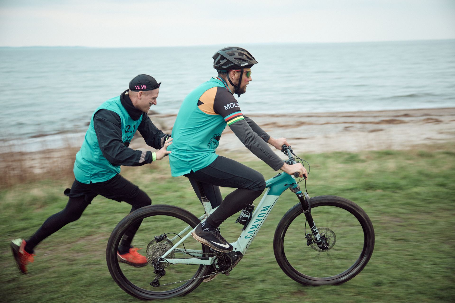 Mand på cykel bliver skubbet af en anden mand på en græsklædt strand.