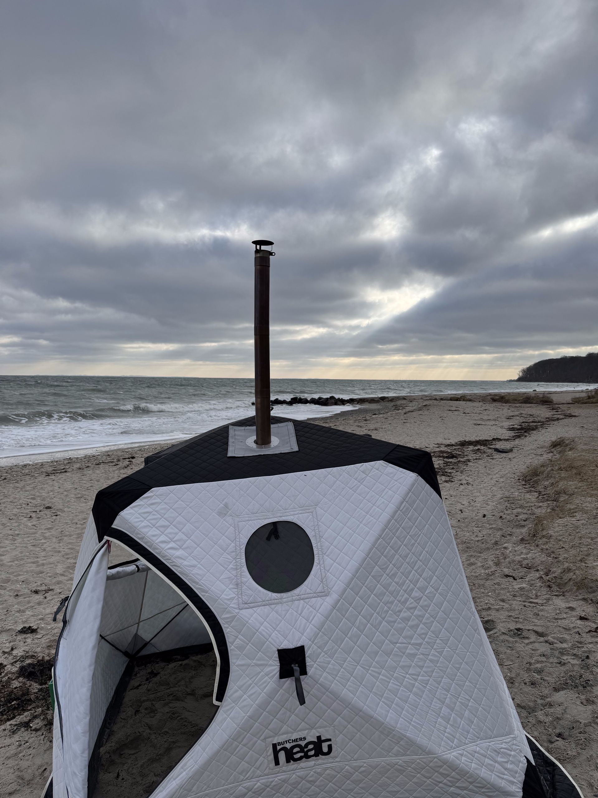 Et lille saunatelt med skorsten på en sandstrand, ud mod havet under en overskyet himmel.