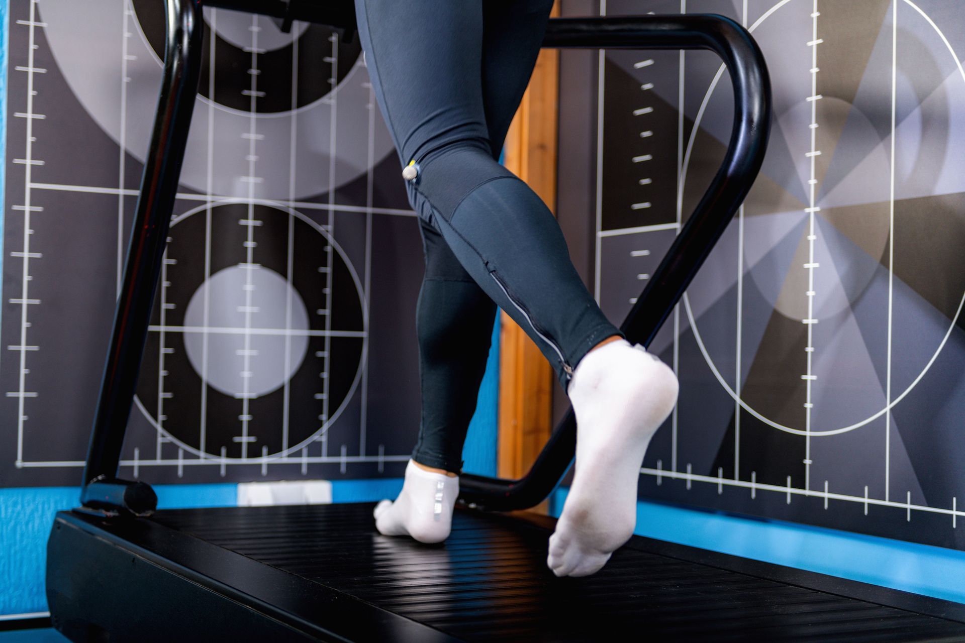 Person's legs in black leggings and white socks walking on a treadmill, against a target-patterned wall.