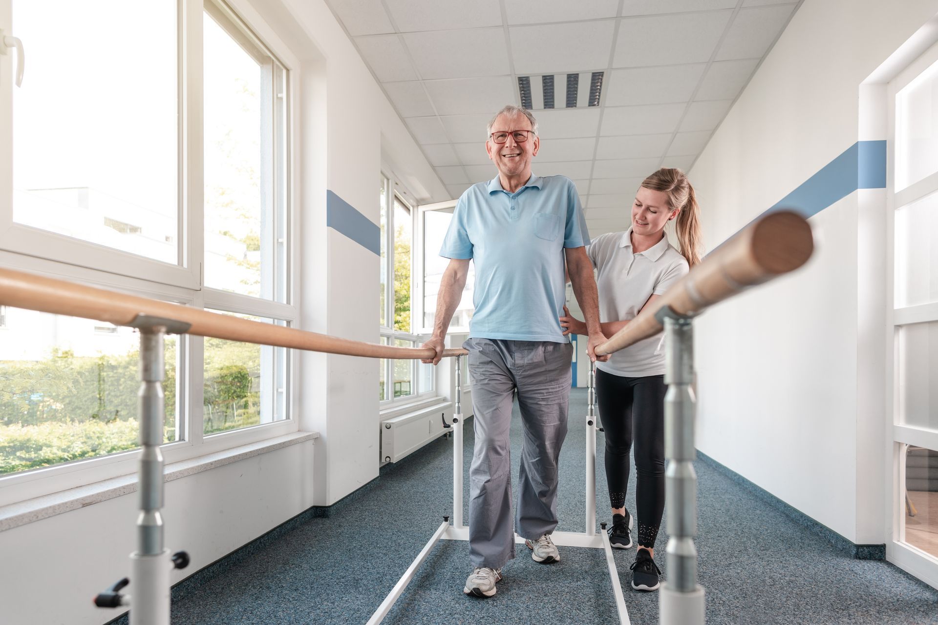 Man walking in rehab facility with therapist, using parallel bars for support.