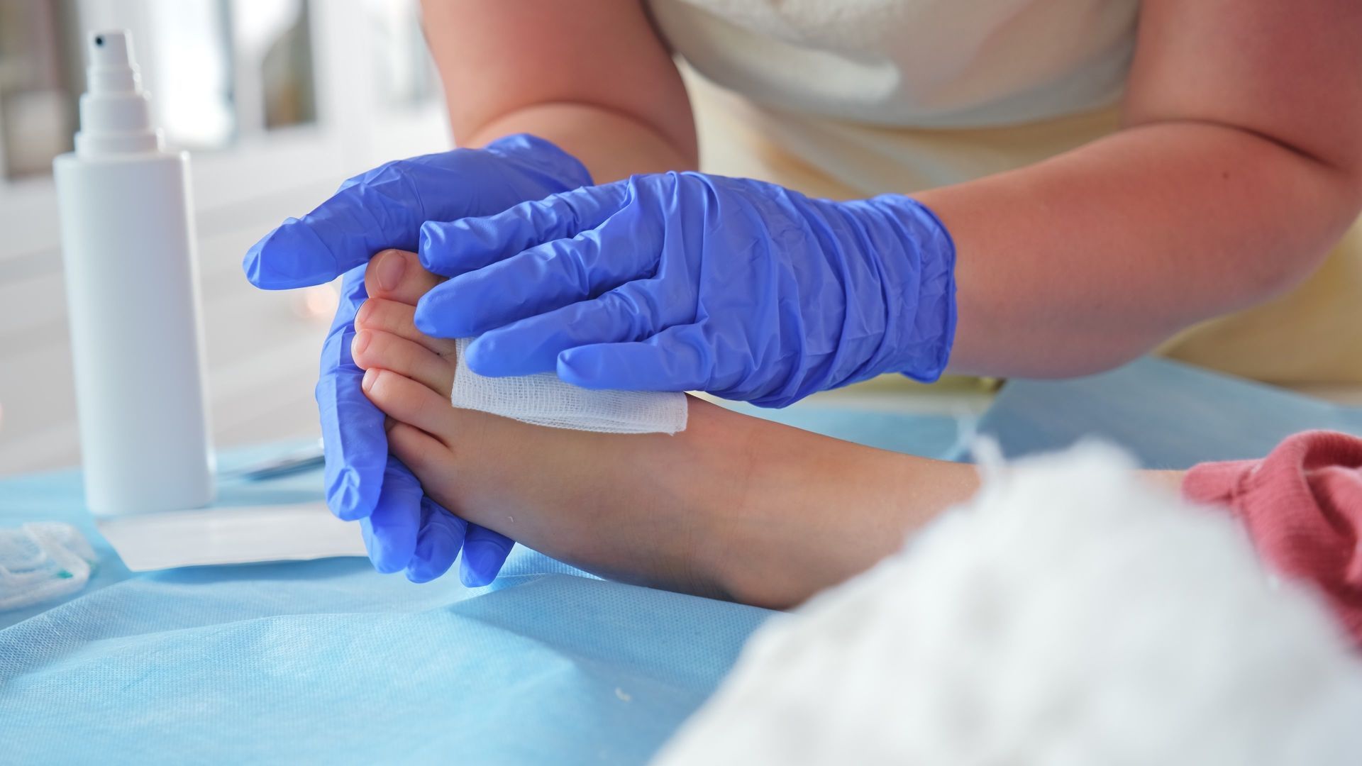 Hands in blue gloves holding a gauze pad on a foot on a blue surface, a bottle in the background.