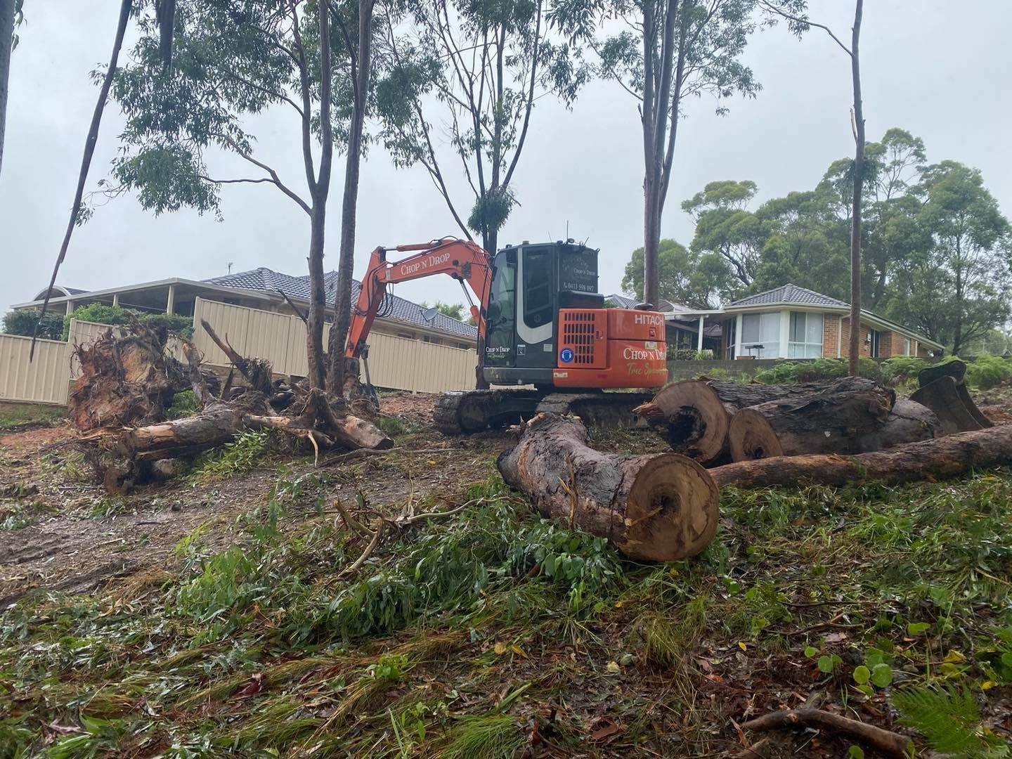 An Excavator Clearing Trees Near Houses, Logs and Debris on the Ground — Chop n Drop Tree Specialists in Valentine, NSW