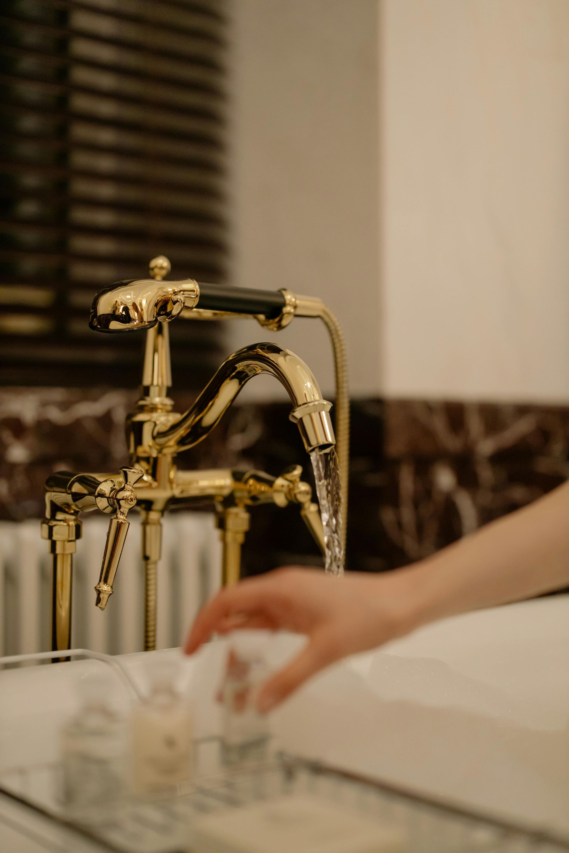 Gold faucet with water running into a bathtub. A hand reaches for toiletries on a tray.