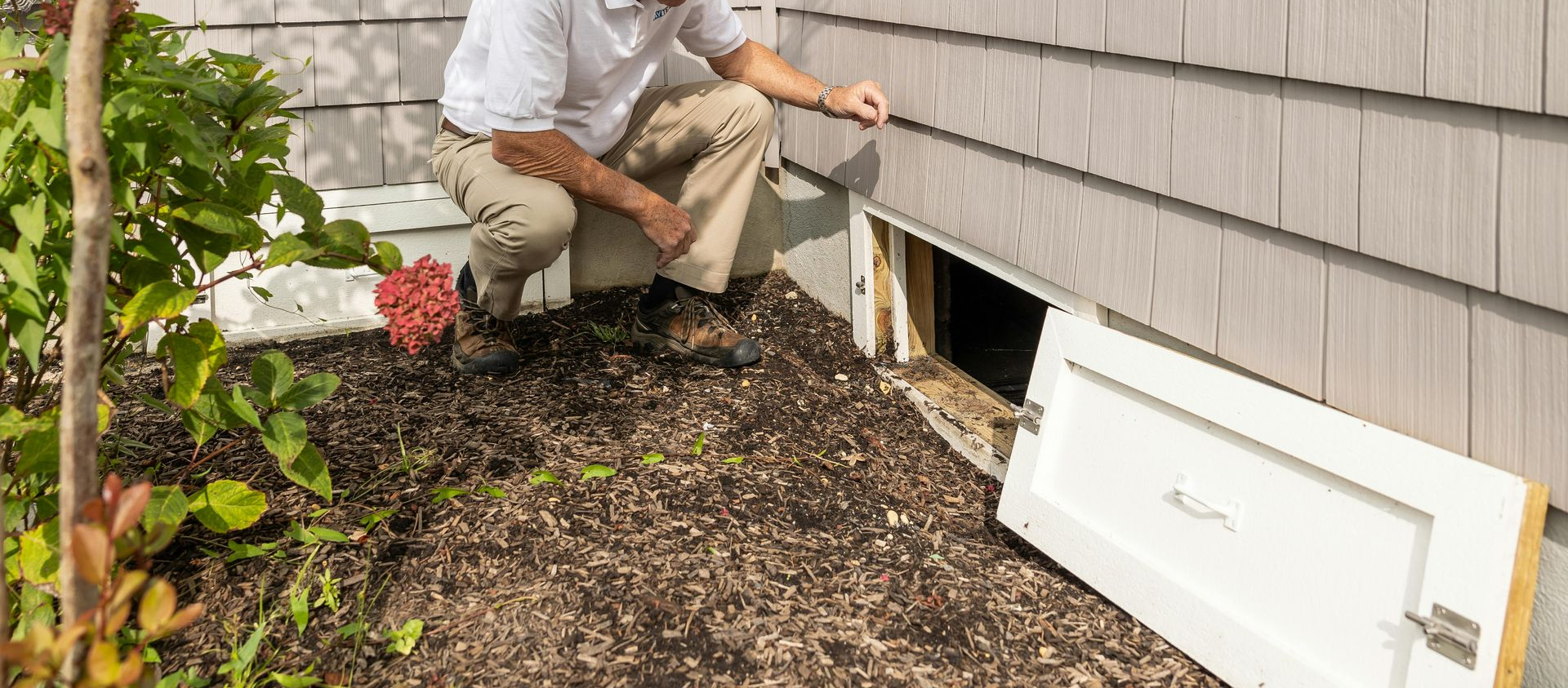 Man crouches near a basement window. The white door is open, and he points at it. Outdoors with mulch and plants.
