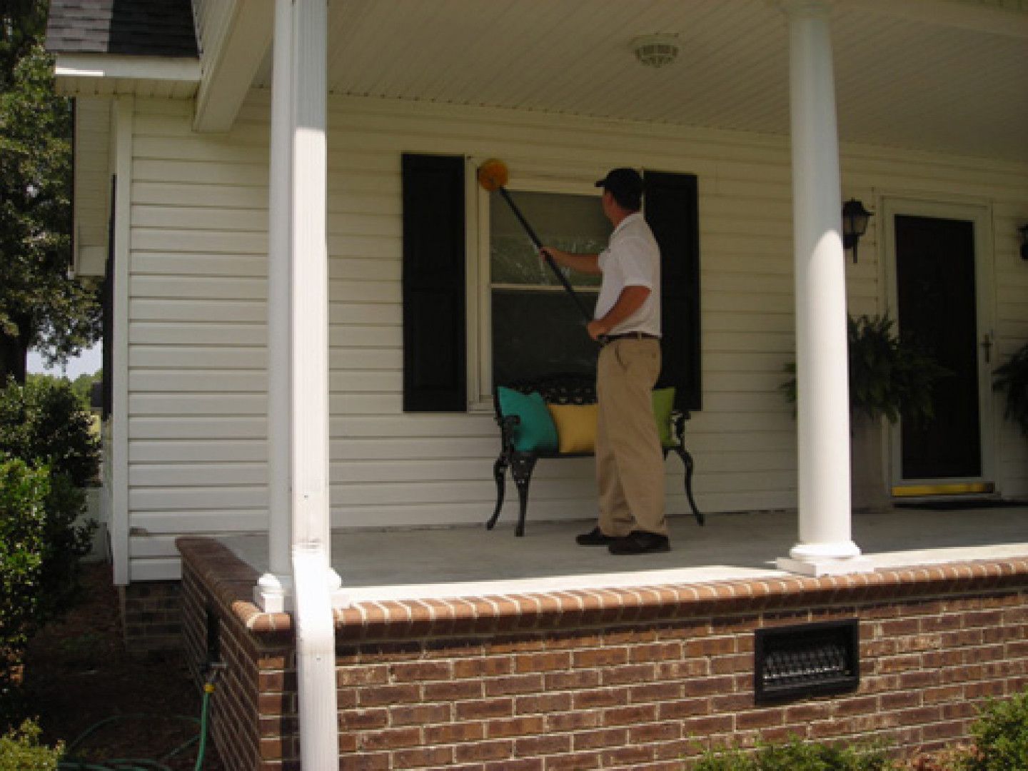 Man cleaning a window from a porch using a long-handled tool. White house with dark shutters and brickwork.