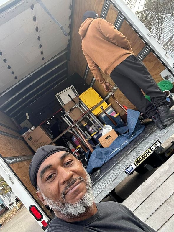 A man in a black skullcap smiles for a selfie inside a moving truck, with another person loading boxes in the background.