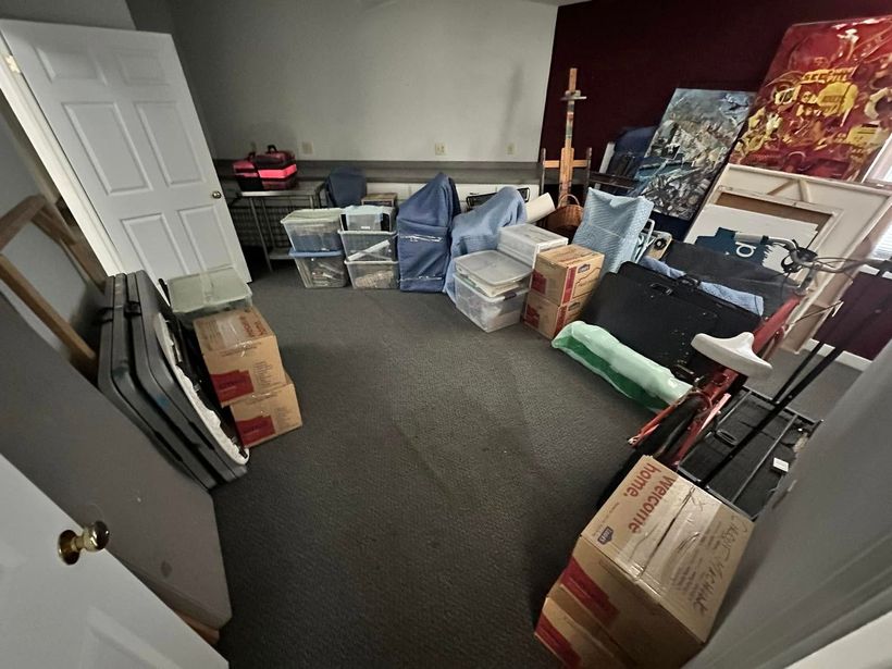 A room filled with stacked cardboard boxes, folded tables, and various storage bins on a gray carpeted floor.