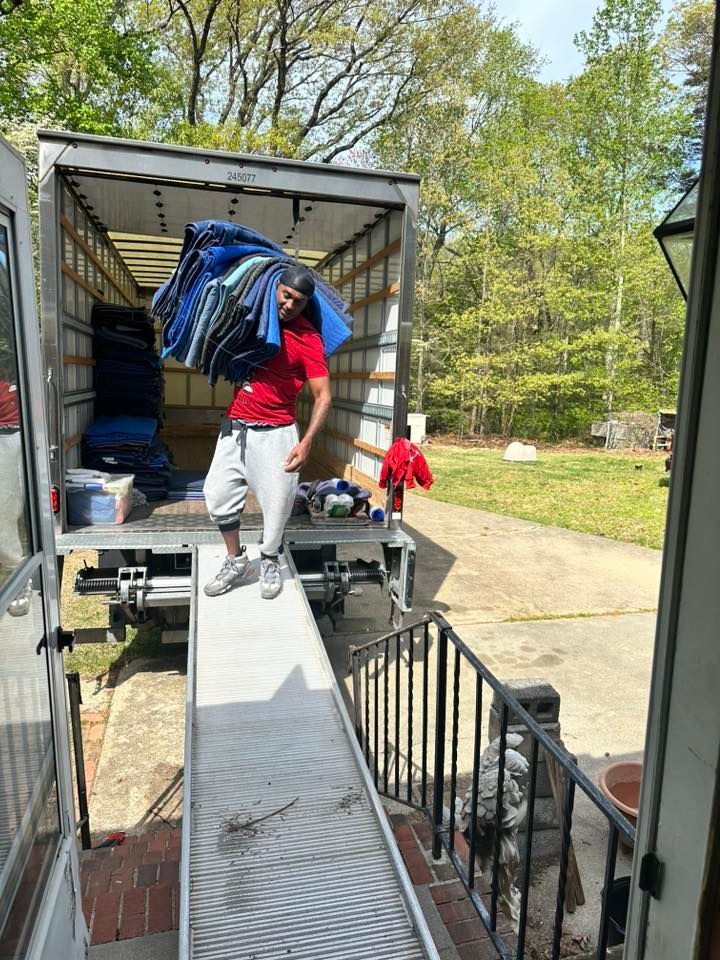 A person in a red shirt and gray sweatpants walks down a moving truck ramp, carrying a stack of blue moving blankets.