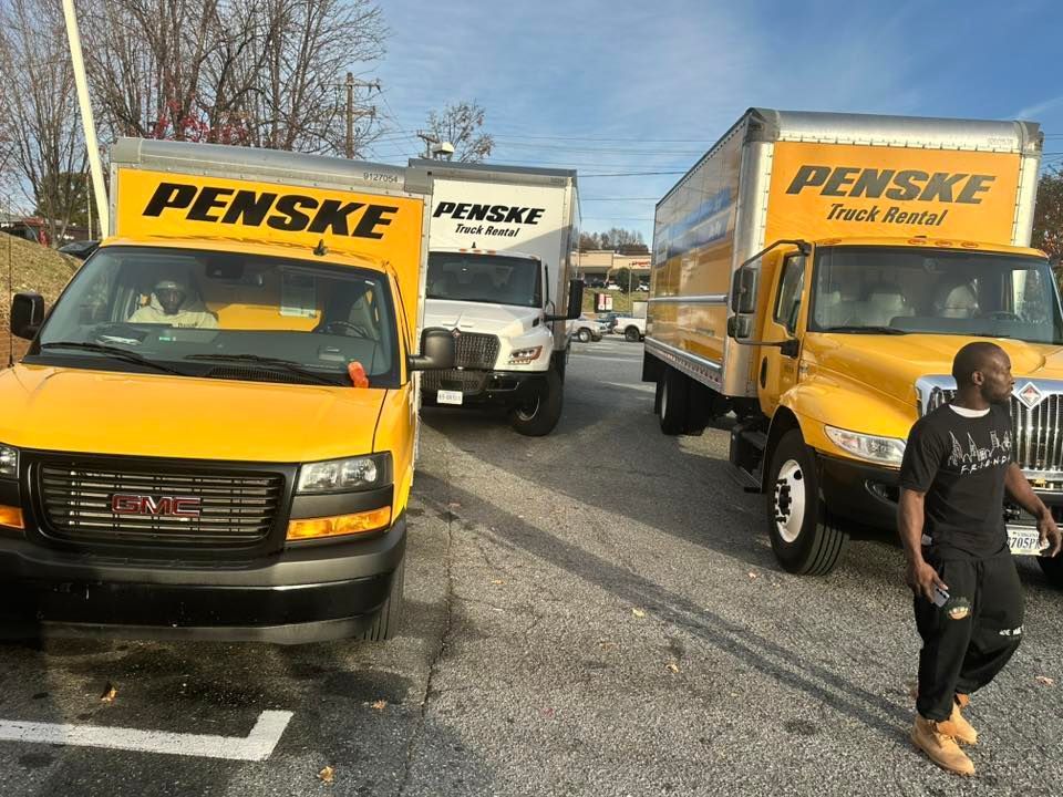 Three yellow Penske rental trucks parked in a gravel lot with a person walking in the foreground.
