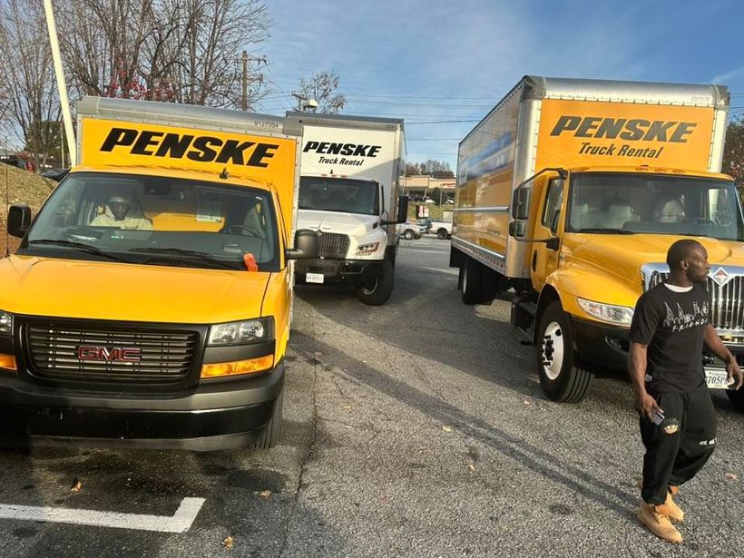 Three yellow Penske rental trucks parked in a lot, with a person walking in the foreground.