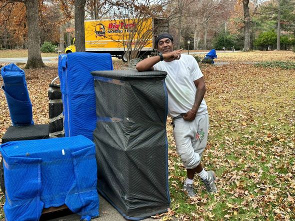 A person leans on a black-wrapped object outdoors near a moving truck, with blue-wrapped items and fall leaves nearby.