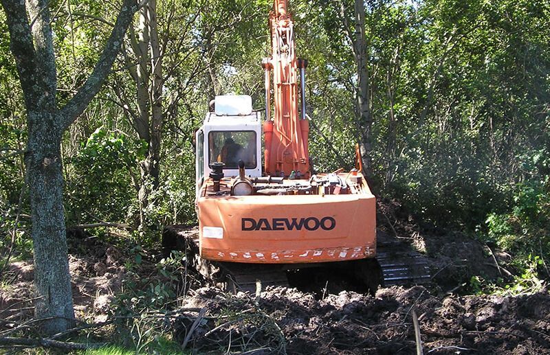 Una excavadora Daewoo de color naranja se encuentra en una zona boscosa, rodeada de tierra removida y árboles.