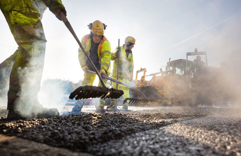 Obreros de la construcción, equipados con ropa de alta visibilidad, rastrillan asfalto humeante en una obra vial.