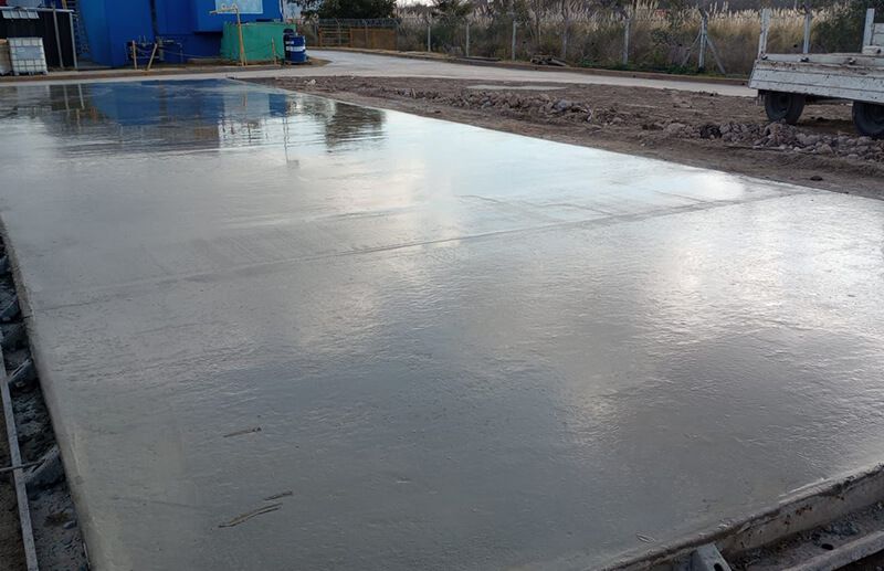 Una losa de hormigón recién vertida y húmeda al aire libre, enmarcada por tablones de madera, con una estructura azul al fondo.