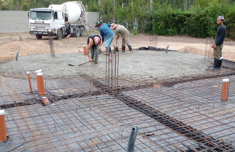Obreros de la construcción nivelan el hormigón fresco en un solar de cimentación, con un camión hormigonera al fondo.