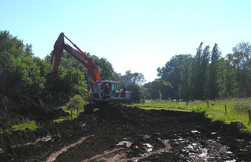 Una excavadora naranja trabaja en un campo fangoso con árboles al fondo en un día soleado.