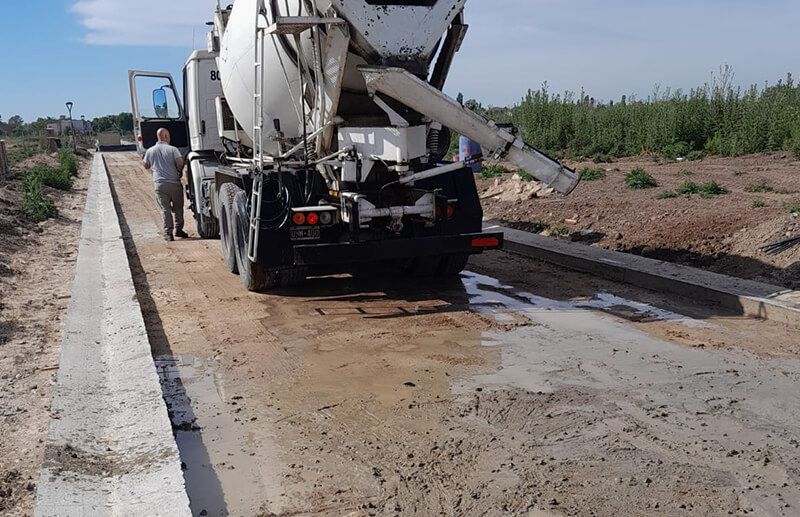 Un camión hormigonera vierte hormigón fresco sobre un camino de tierra entre bordillos de hormigón recién instalados en una obra en construcción.