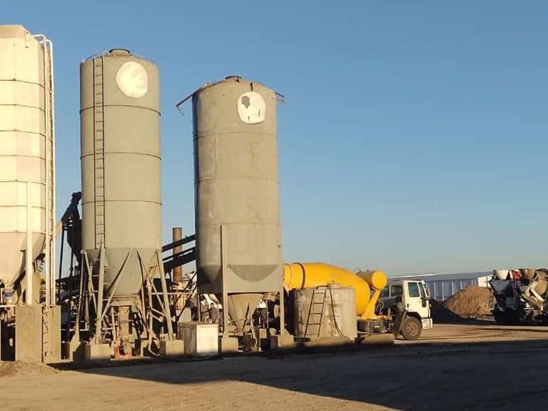 Una planta de hormigón con grandes silos industriales grises y un camión hormigonera amarillo aparcado en un patio de grava.