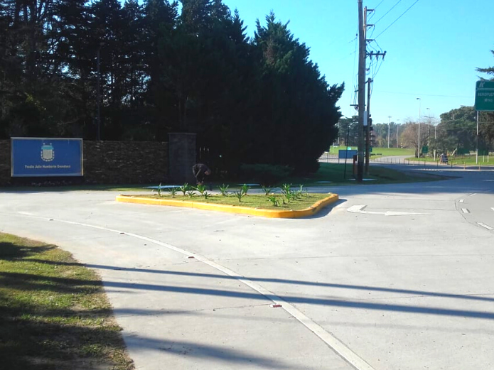Una entrada con muro de piedra y un letrero azul, una mediana triangular ajardinada y un poste de servicios públicos en una carretera soleada.