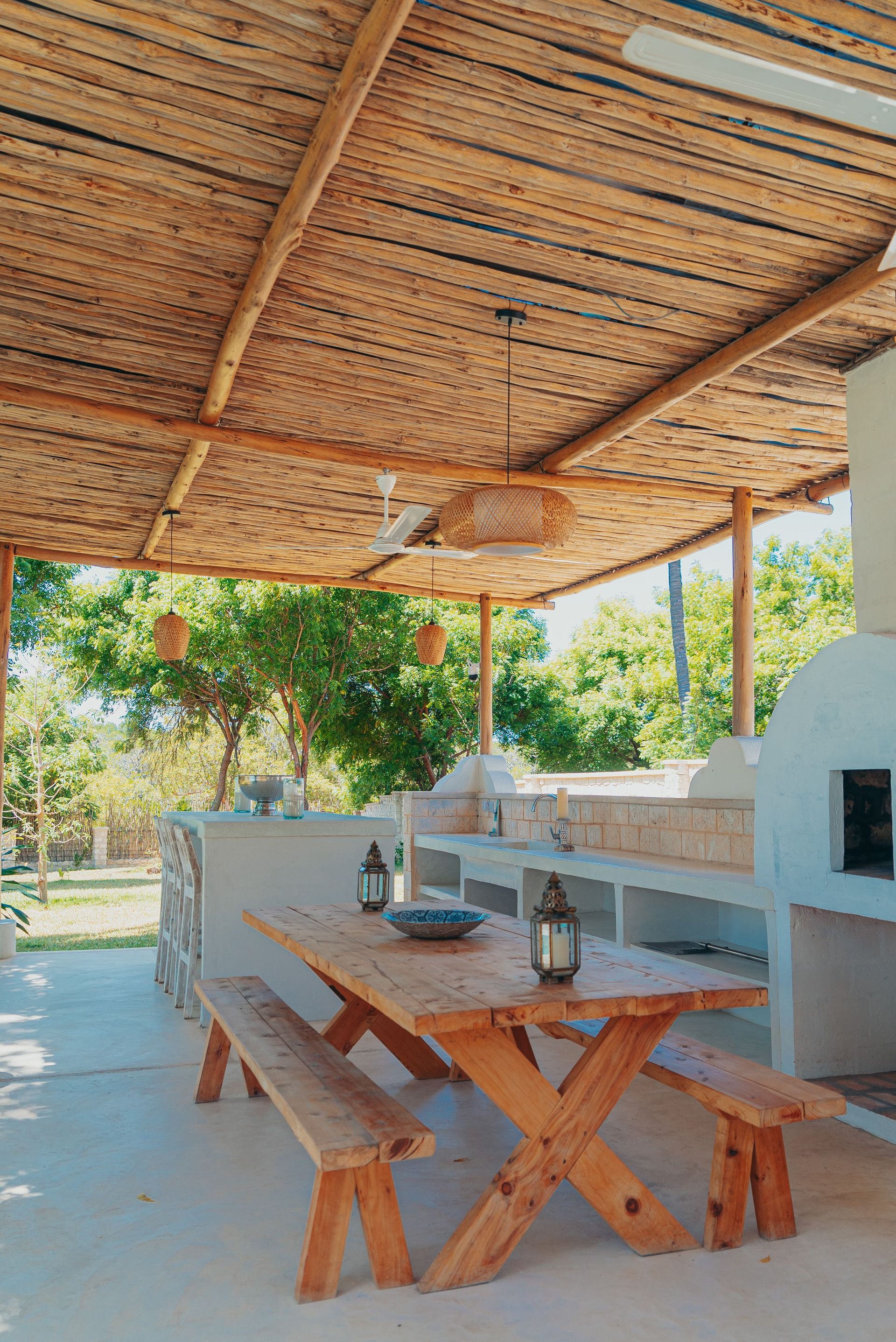 Rustic outdoor dining area with wooden table and benches under a woven shade roof, beside a white grill.