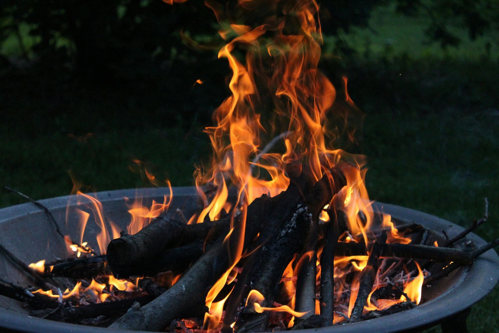 Burning campfire in a metal fire pit at dusk, with orange flames and logs against a dark outdoor background