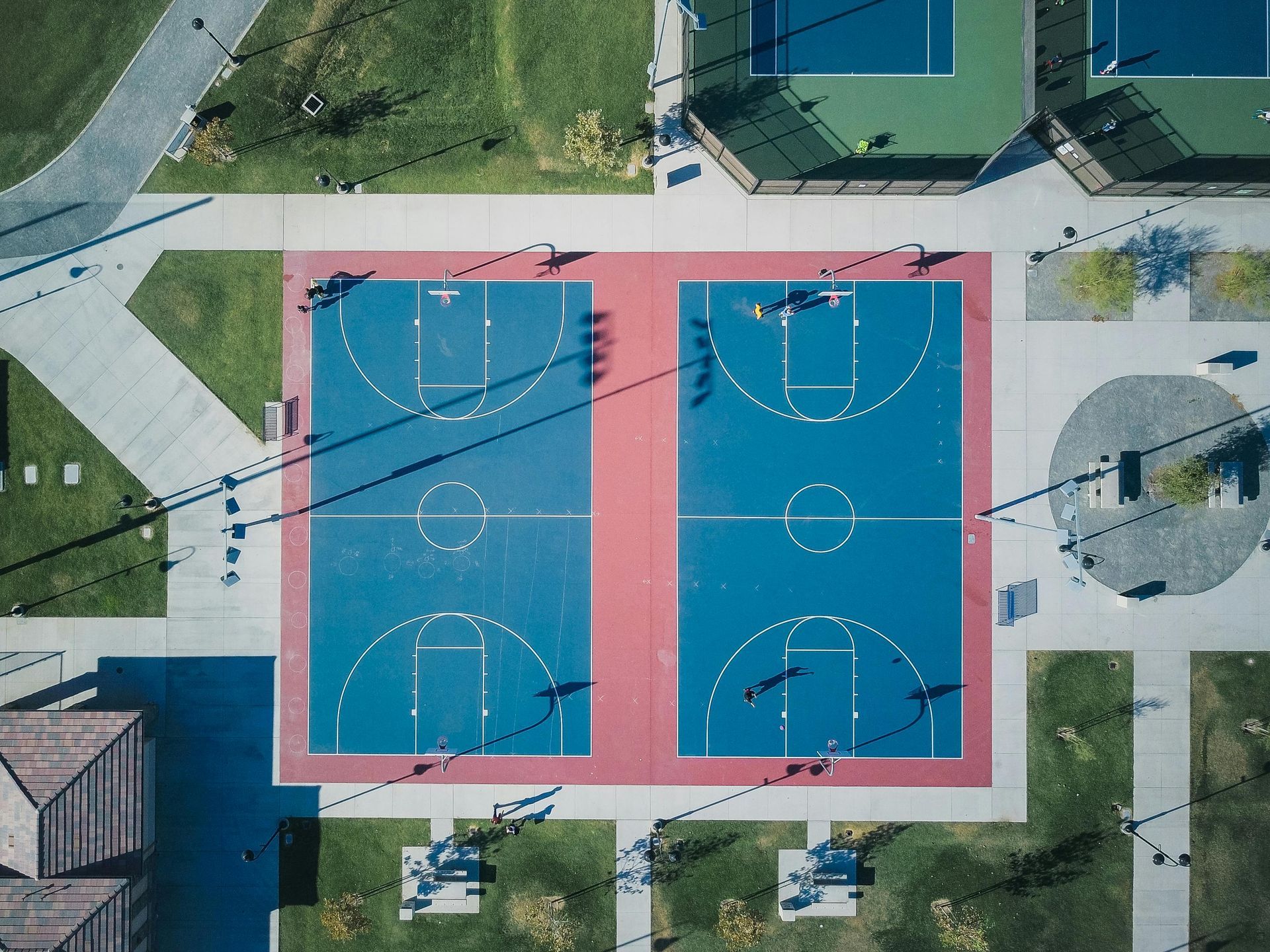 Overhead view of two blue basketball courts with players. Red borders, concrete pathways, and tennis courts nearby.
