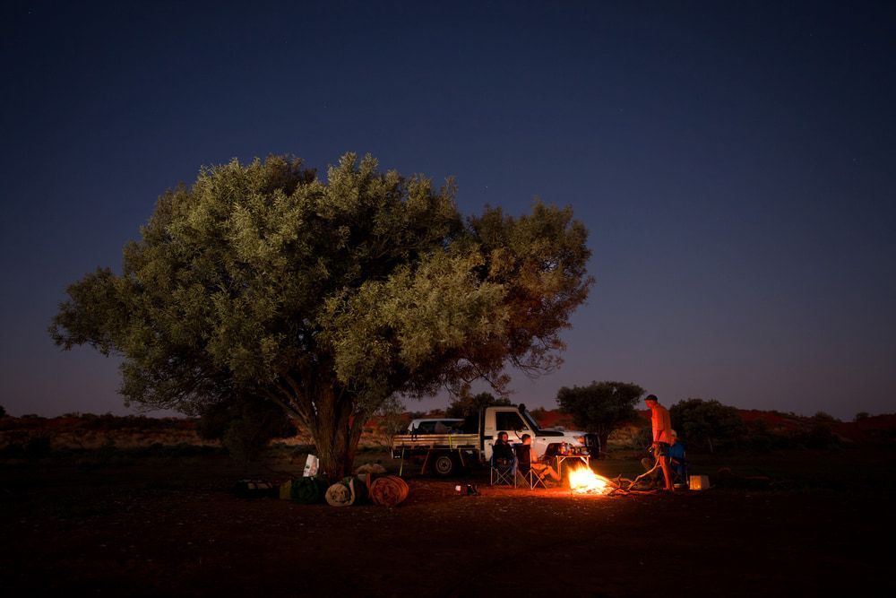 A Group Of People Are Standing Around A Campfire Under A Tree — PH Davie in Boulia, QLD