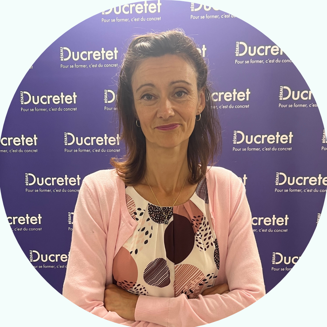 Femme, bras croisés, devant un fond violet orné des logos Ducretet. Elle porte un cardigan rose et un haut à motifs.