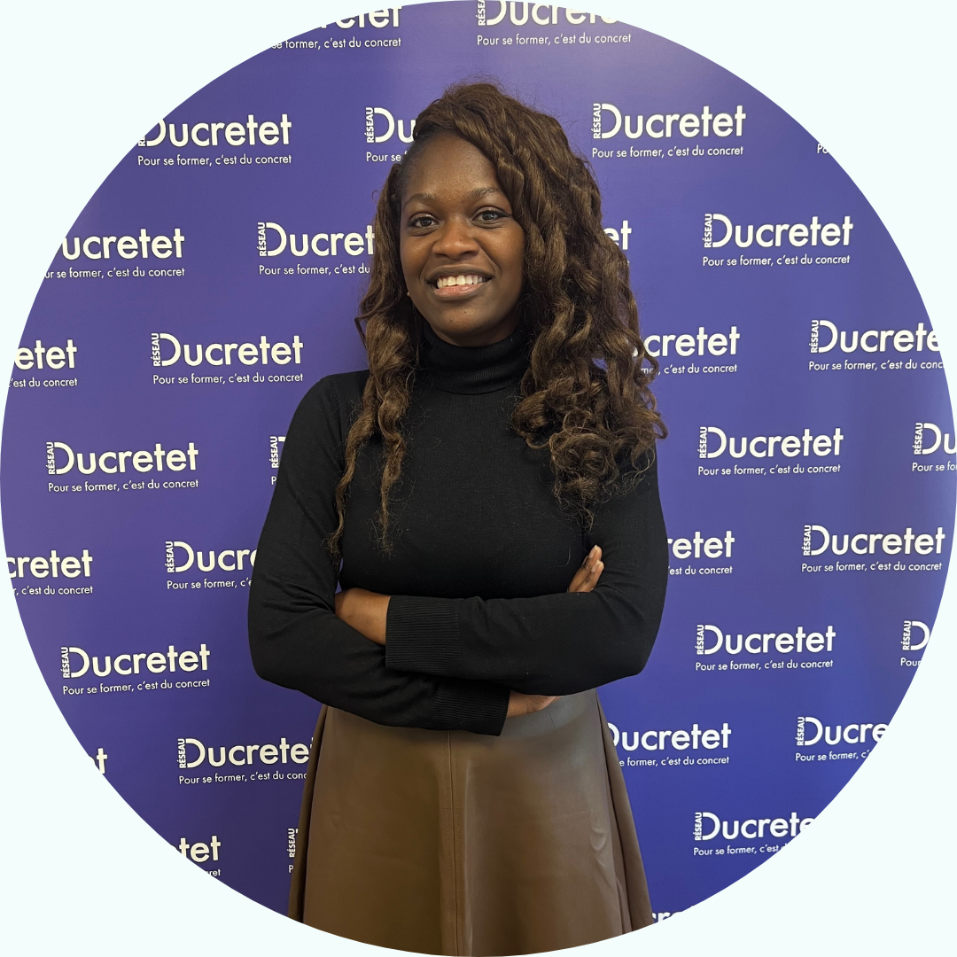 Femme aux bras croisés, souriante, vêtue d'un col roulé noir et d'une jupe marron ; devant un fond violet Ducretet.