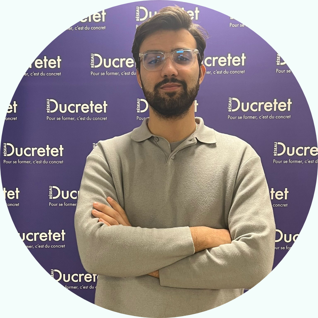 Un homme portant des lunettes et une barbe, les bras croisés, se tient devant un fond de scène avec le logo « Ducretet ».