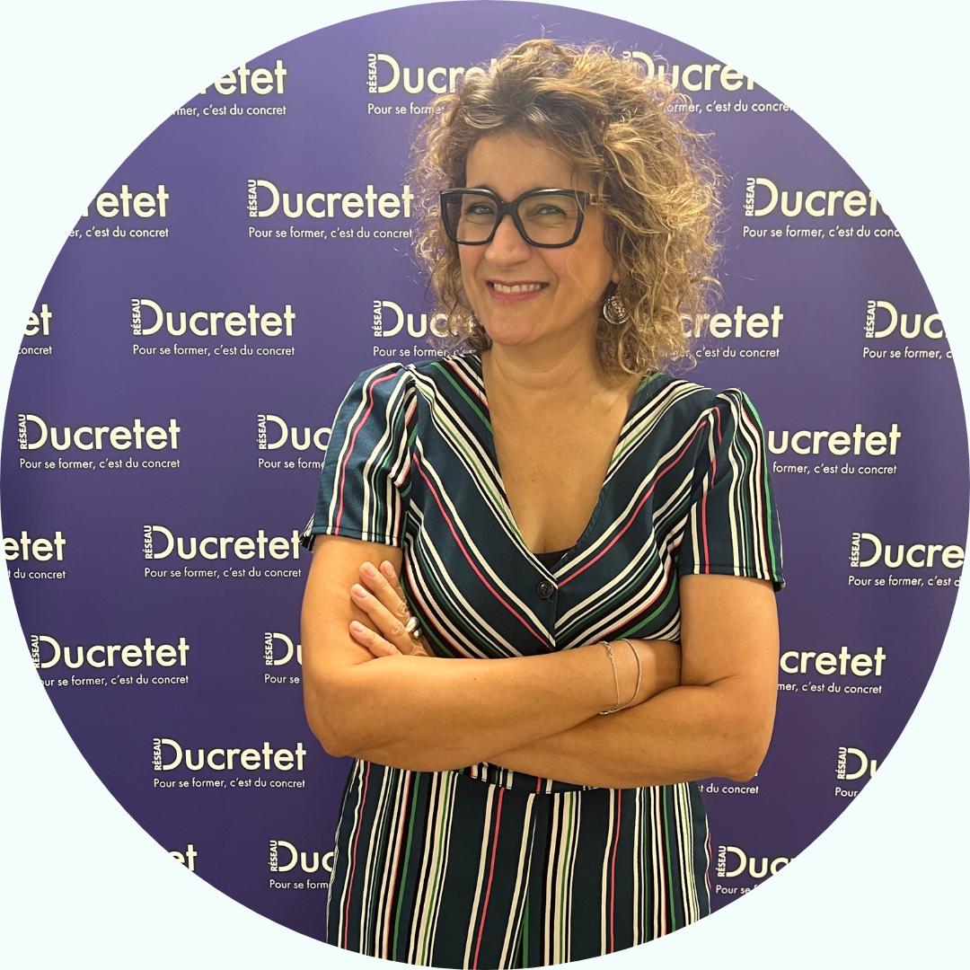 Femme aux cheveux bouclés portant des lunettes, robe rayée, bras croisés, souriant devant un fond de marque 