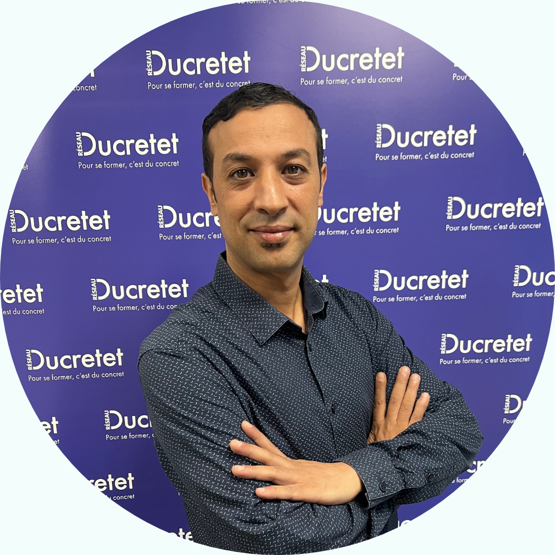 Homme aux bras croisés devant un décor Ducretet. Il a les cheveux noirs courts et un léger sourire.