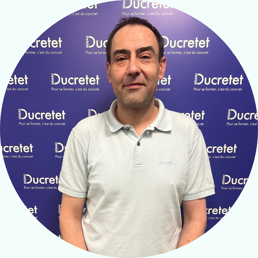 Homme portant un polo de couleur claire, debout devant un mur portant la marque « Ducretet » répétée.