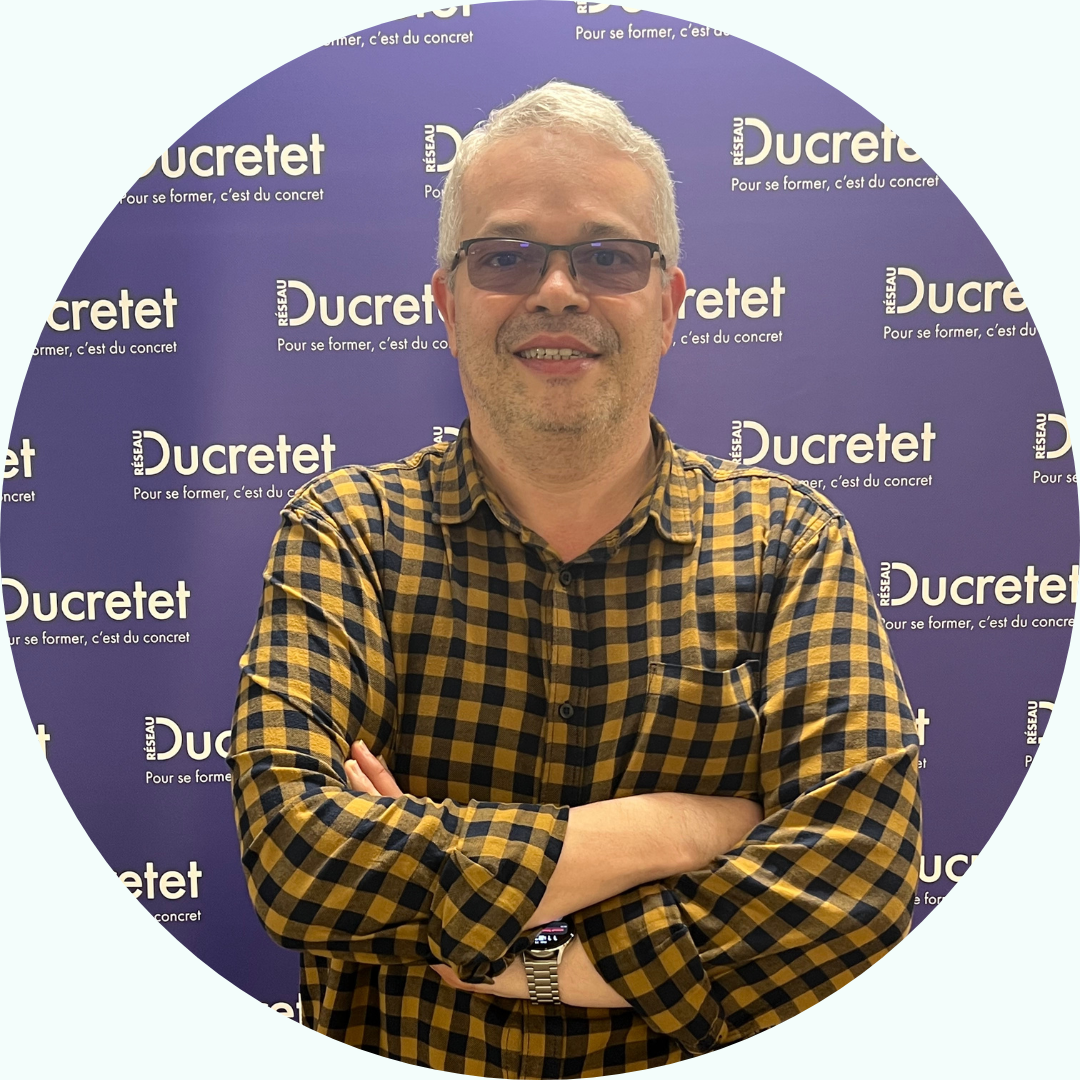 Homme aux cheveux gris et lunettes en chemise à carreaux jaune et noir, les bras croisés, devant un fond violet Ducrretet.