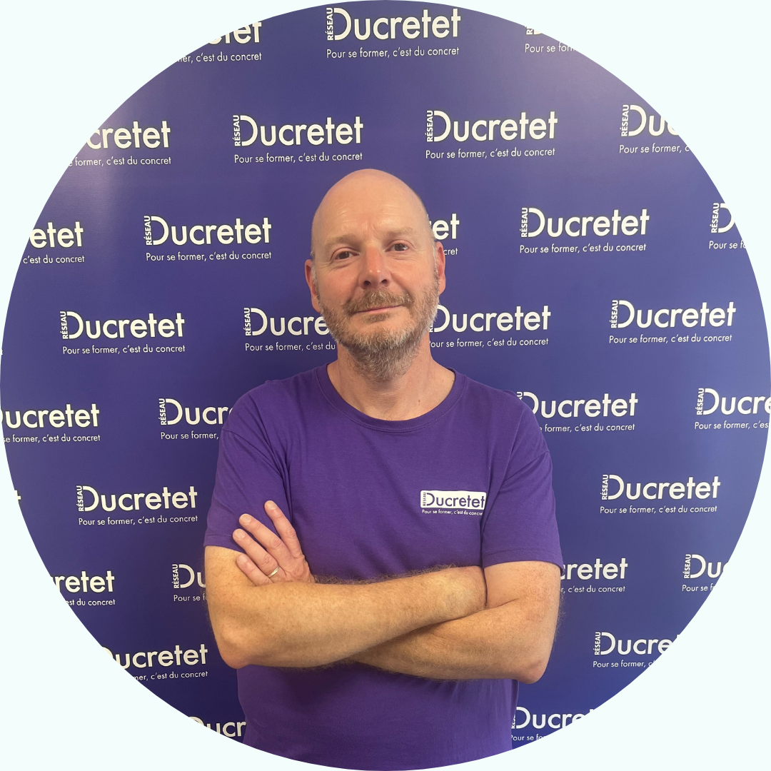 Homme aux bras croisés, portant une chemise violette, debout devant un mur avec « Ducretet » répété.