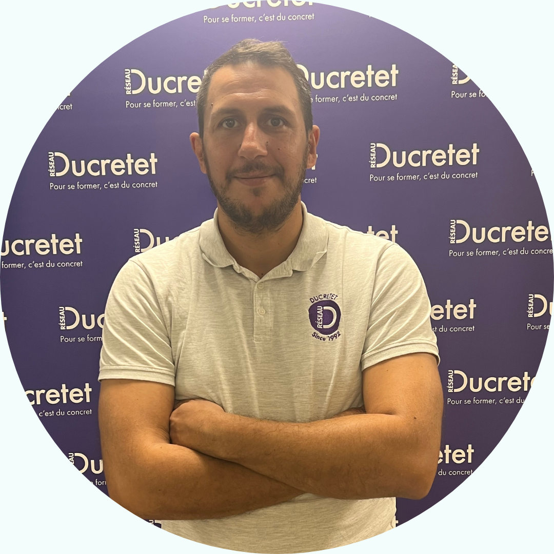 Homme aux bras croisés, vêtu d'un polo gris, debout devant un fond violet avec des logos 