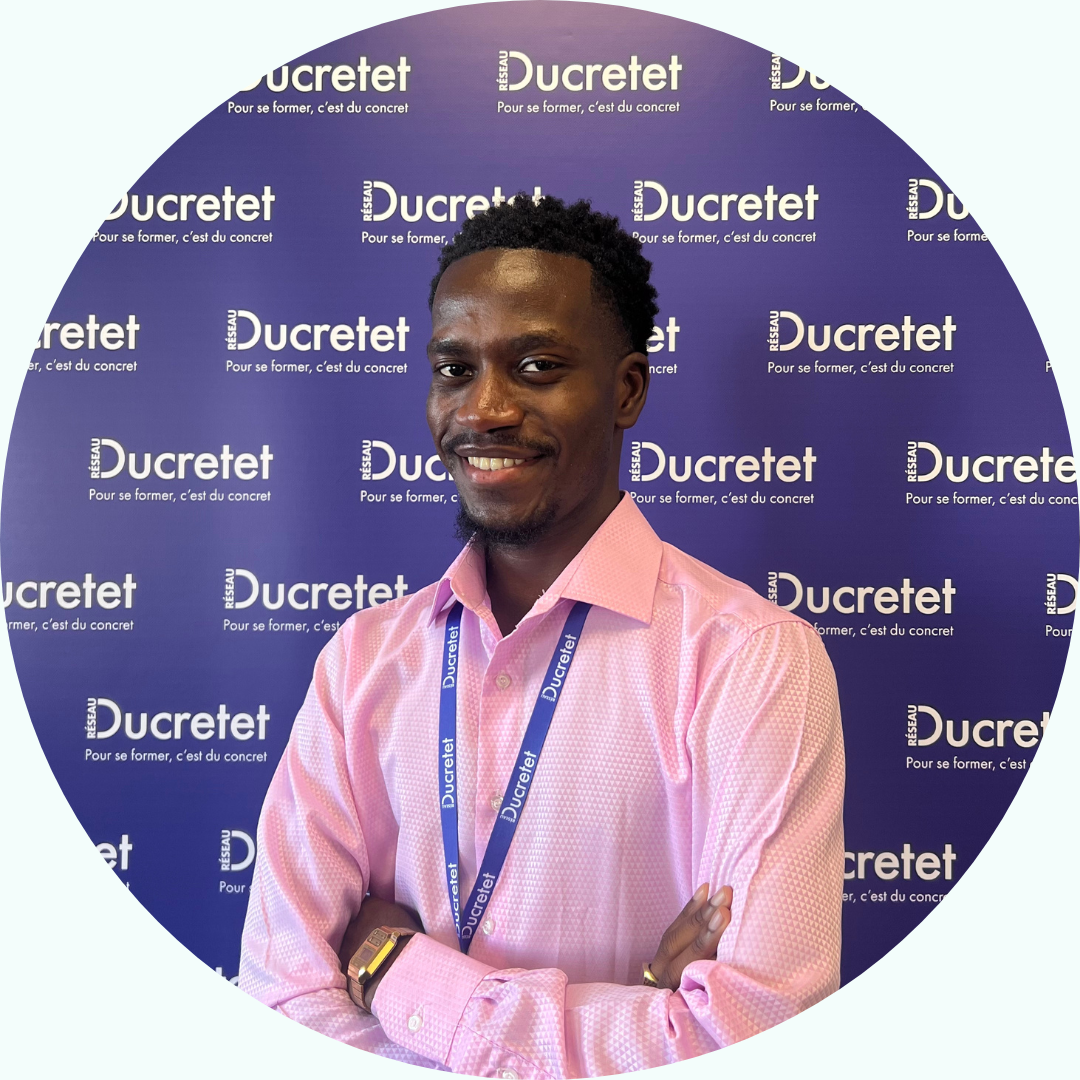 Un homme noir souriant, les bras croisés, portant une chemise rose et un cordon devant un fond « Ducretet ».