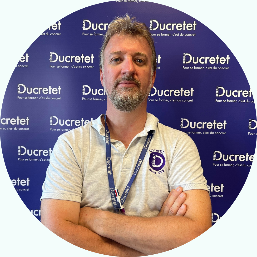 Homme, bras croisés, devant un décor « Ducretet ». Il porte un polo gris et un cordon.