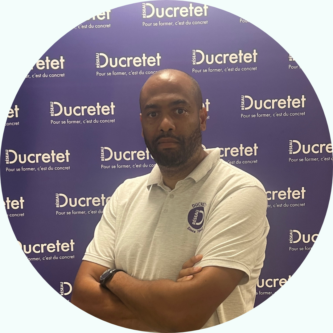Homme aux bras croisés, chemise grise, devant un fond de logo Ducretet.