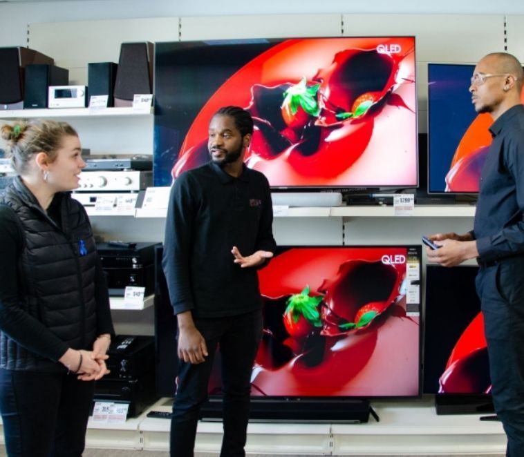 Trois personnes examinent des téléviseurs dans un magasin d'électronique. L'une d'elles fait un geste.