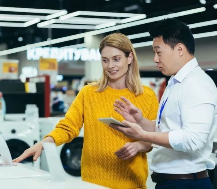 Une femme en pull jaune et employée du magasin observent un présentoir de machines à laver ; l’employée tient une tablette.