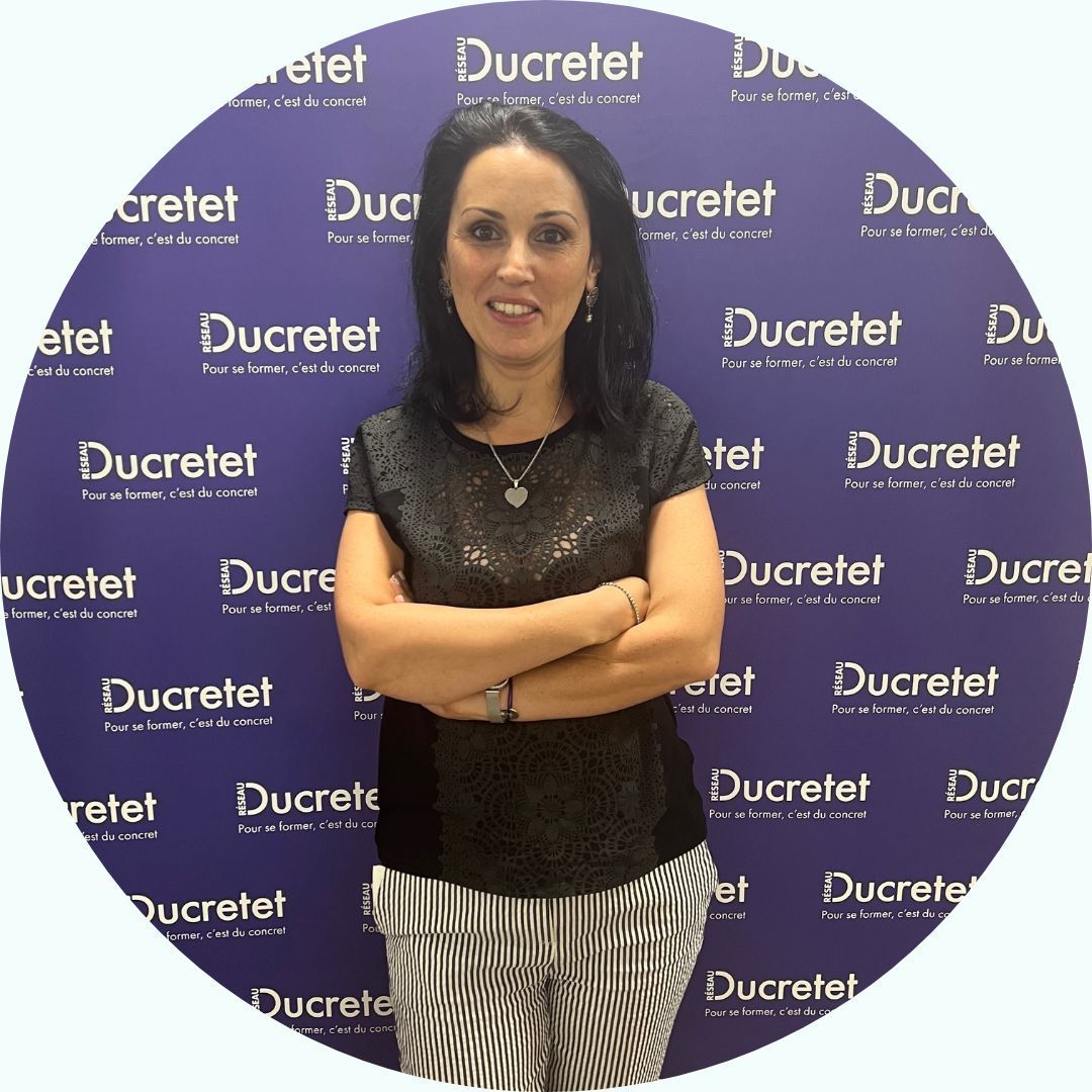 Femme, bras croisés, souriante, devant un fond violet « Ducretet ». Elle porte un haut noir et un pantalon rayé.