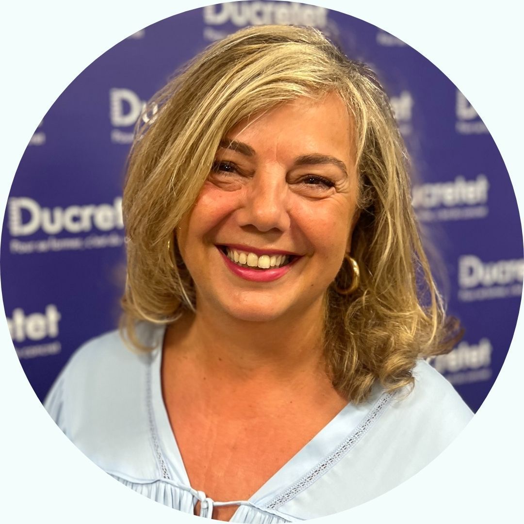 Femme souriante aux cheveux blonds, vêtue d'une chemise bleu clair, devant un logo Ducrêt violet.