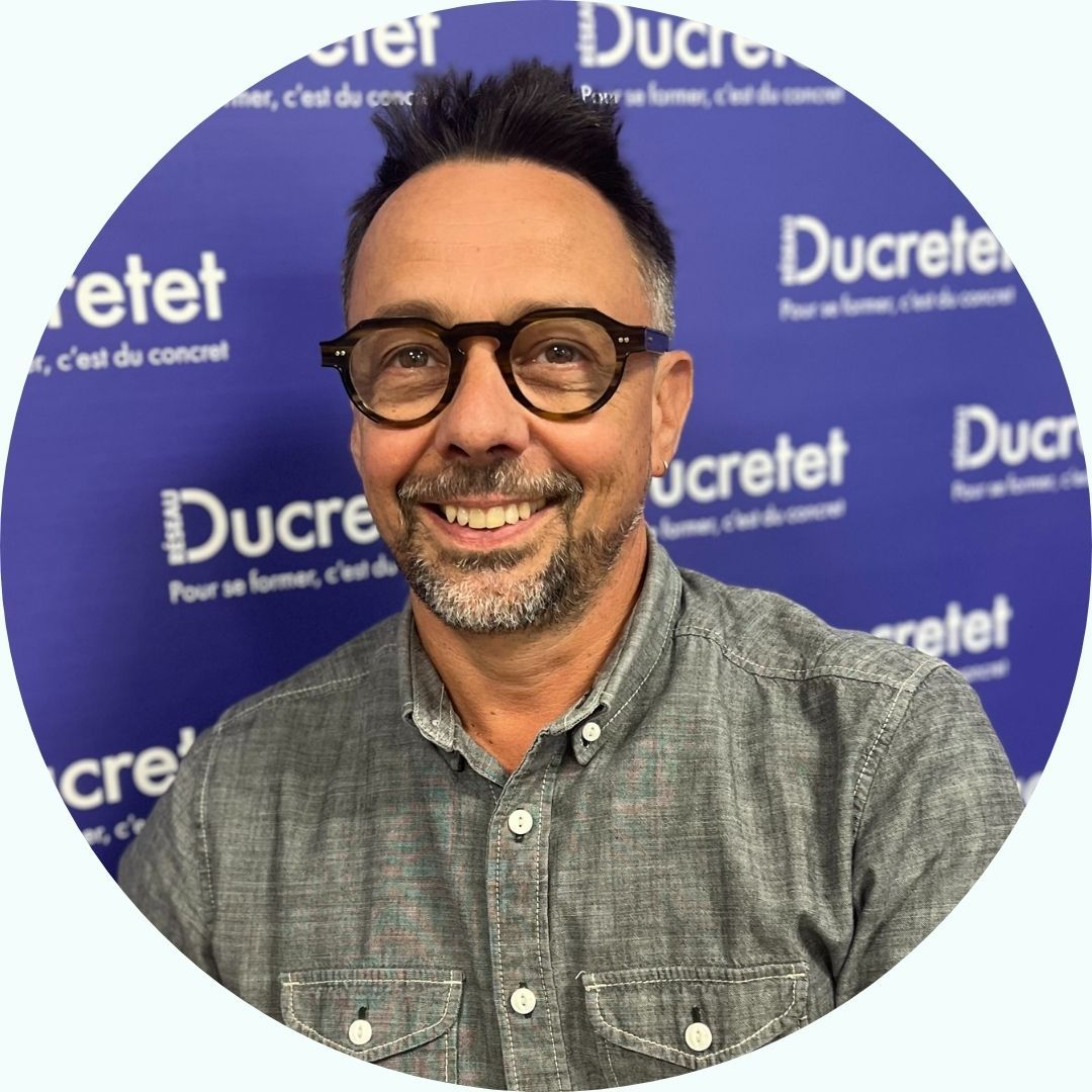 Un homme à lunettes sourit devant un fond bleu sur lequel est répété le mot « Ducretet ».