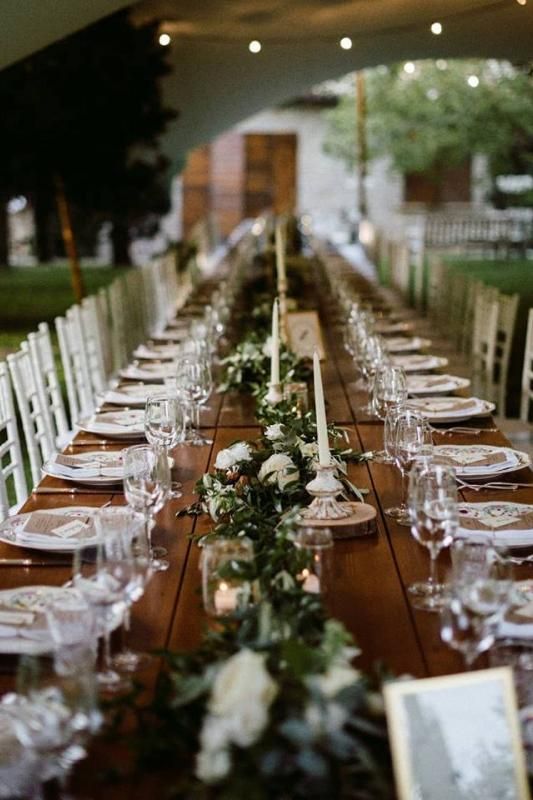Un lungo tavolo di legno con sopra piatti, bicchieri, candele e fiori.