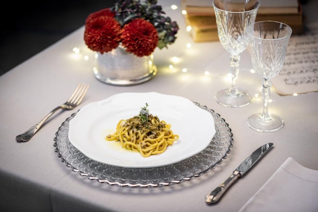 Su un tavolo c'è un piatto di spaghetti accanto a bicchieri di vino e un vaso di fiori.