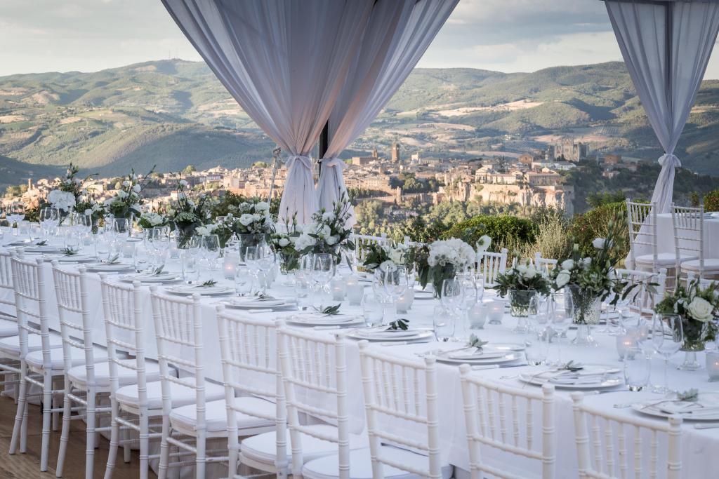 Un lungo tavolo con sedie bianche sotto una tettoia con vista sulla città.