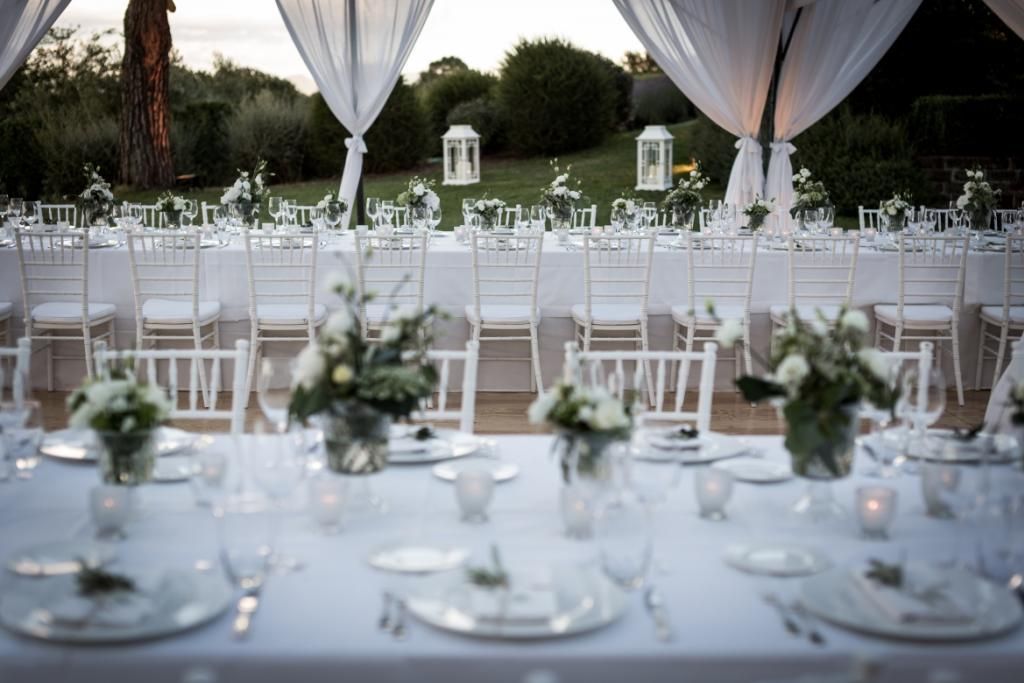 Una lunga tavola con piatti, bicchieri e vasi di fiori.
